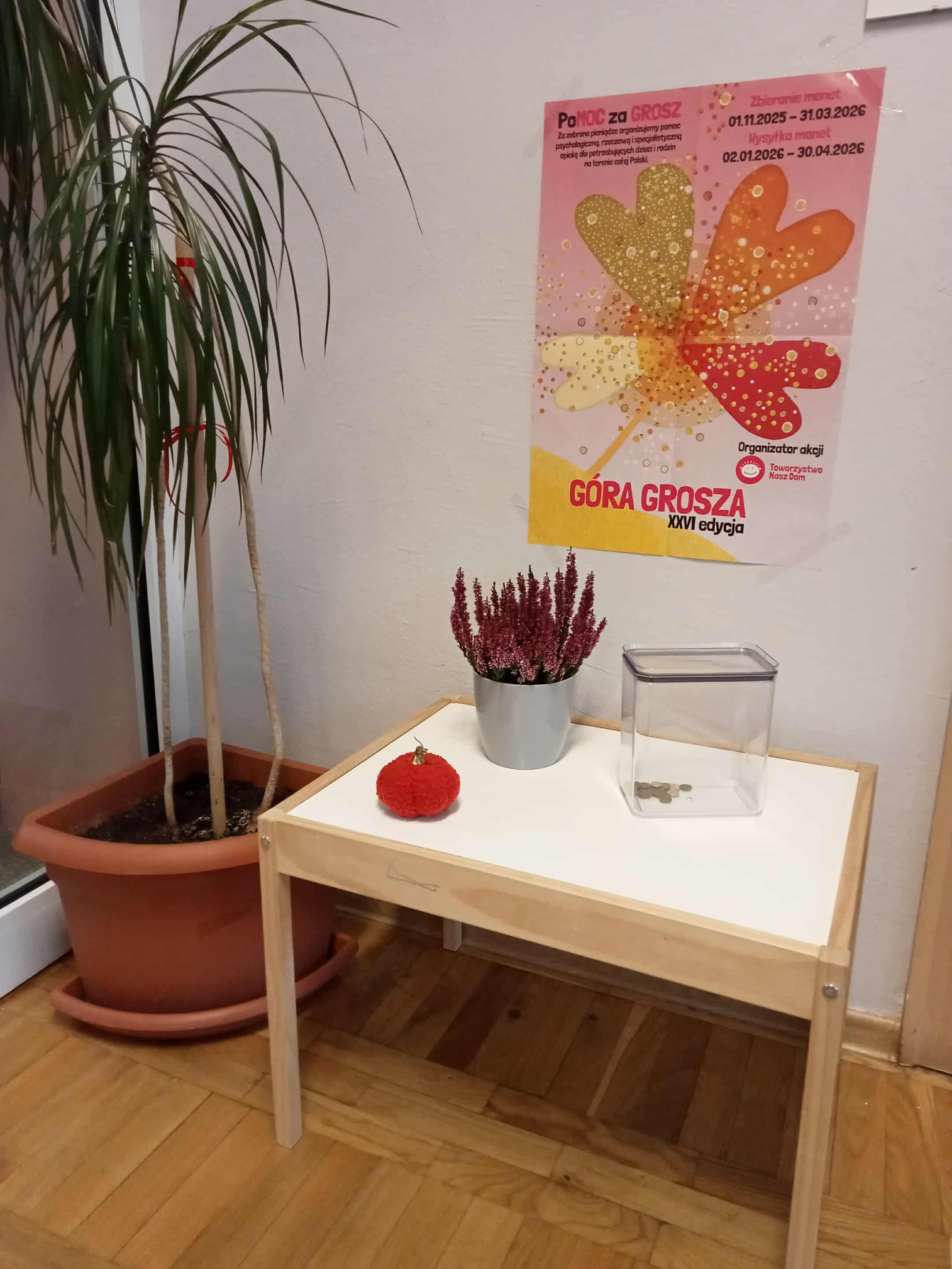Zdjęcie miejsca zbiórki monet. Mały stolik na którym znajduje się puszka na monety. Nad stolikiem przywieszony plakat informacyjny o zbiórce.